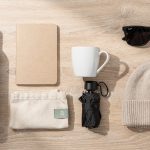 Objets publicitaires saisonniers : adapter ses campagnes Objets publicitaires posés sur un bureau en bois clair : gourde isotherme, carnet, tote bag, mug, parapluie, lunettes de soleil et bonnet