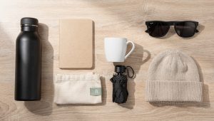 Objets publicitaires posés sur un bureau en bois clair : gourde isotherme, carnet, tote bag, mug, parapluie, lunettes de soleil et bonnet